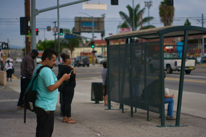 Erick Huerta checks his phone as he waits for the bus on Western Ave. at Exposition Blvd. At this point he has already taken one bus and one train and has been in transit for an hour and fifteen minutes. Sahra Sulaiman/Streetsblog L.A.